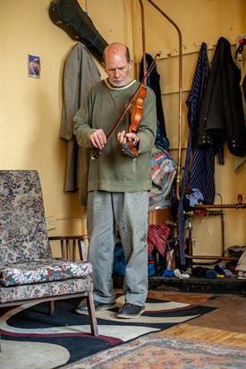 An elderly man in faded clothes stands in a room of his apartment and plays a violin.